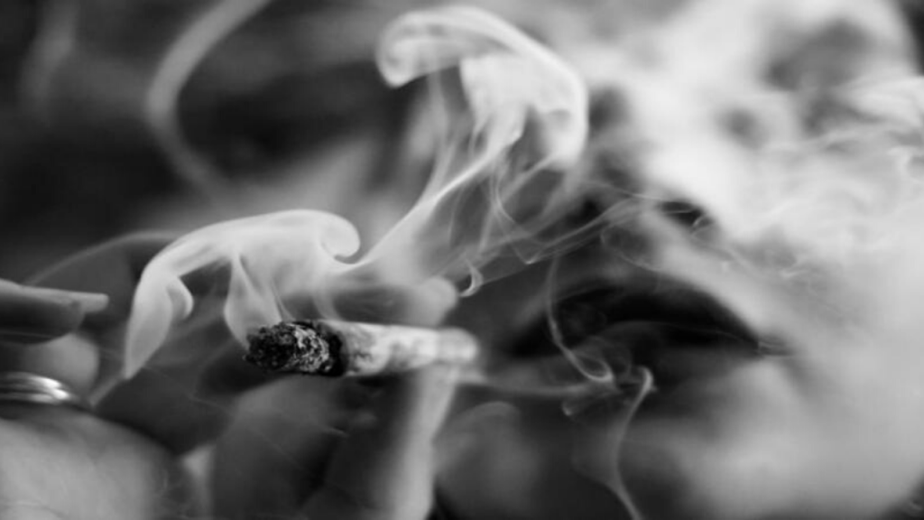 Close up image of a cigarette with smoke and a person's mouth in the background.