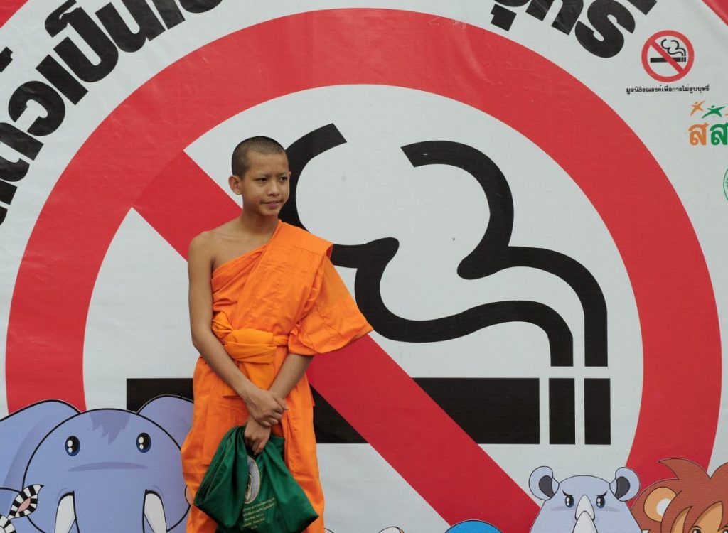 Monk in Thailand wearing orange robe standing in front of an anti-smoking banner.